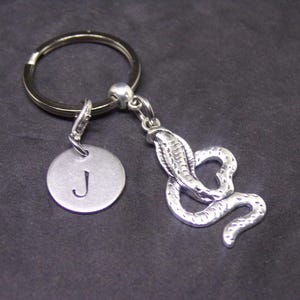 May include: Silver-coloured keyring featuring a cobra pendant and a round charm with the letter "J". The cobra is detailed with scales and a raised hood. The keyring and charms are made of metal.