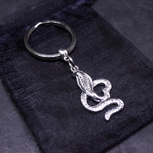 May include: A silver-toned metal keyring featuring a detailed cobra snake pendant with textured scales. The snake is coiled in a curved position and is attached to a standard circular split ring, providing a functional accessory for keys.