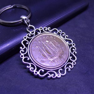 May include: A silver-toned keychain featuring a vintage coin with a ship design. The coin is set within an ornate, filigree-style frame. The keychain has a circular ring for attaching keys.