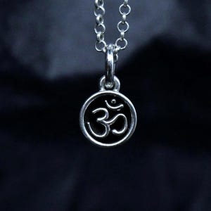 以下が含まれることがあります： 小さな丸いペンダントにオームのシンボルが付いたシルバーカラーのネックレス。ペンダントは繊細なチェーンから吊り下げられています。背景は暗く、質感のある生地です。