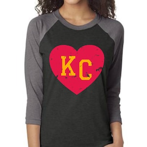 May include: A black and grey raglan t-shirt with a red heart graphic that says "KC" in yellow letters.