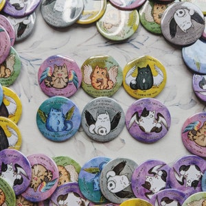 Peut inclure: Plusieurs boutons ronds colorés avec des illustrations de chats ailés. Chaque bouton présente un design et un texte uniques, avec des couleurs variées comme le jaune, le bleu et le violet. Les boutons sont dispersés sur une surface à motifs.