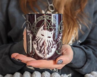 Könnte beinhalten: Eine schwarze Keramiktasse mit einer weißen Katze mit langem Bart und einem Pentagramm-Design. Die Katze ist von einem Kreis mit Runen umgeben. Die Tasse wird von einer Person gehalten.