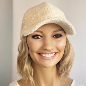 Op de afbeelding: Een vrouw draagt een witte, pluche baseballpet met een gebogen klep. Ze glimlacht en heeft blond haar.