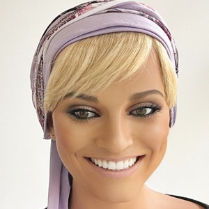 May include: A woman with blonde hair is wearing a purple and white patterned headscarf. The scarf is tied in a knot at the back of her neck and the ends are hanging down her back. She is smiling and looking at the camera.