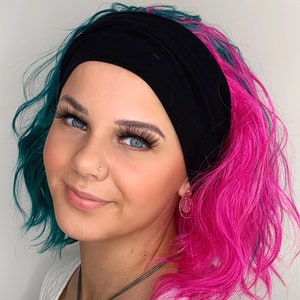 May include: A woman with teal and pink hair is wearing a black headband. She is wearing a white shirt and a silver necklace with a pendant. She is also wearing a gold earring.