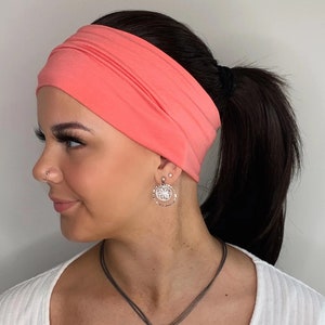May include: A coral pink headband made of soft fabric. The headband is worn by a person with long dark hair pulled back in a ponytail. The person is wearing a white shirt and a silver necklace with a round pendant.