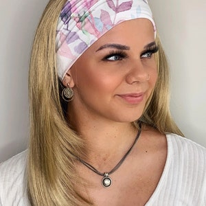 May include: A woman with long blonde hair is wearing a white headband with a floral pattern. The headband features pink, purple, and blue flowers and leaves. She is also wearing a white shirt and a silver necklace with a round pendant.