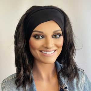 May include: A woman with long, dark brown wavy hair is wearing a black headband. The headband is made of a soft, stretchy fabric and is worn across the forehead.