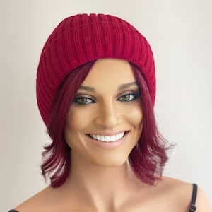 May include: A woman with short, curly, burgundy hair is wearing a red knit beanie. She is smiling and looking at the camera.