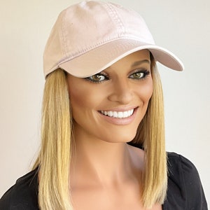 Puede incluir: Una gorra de béisbol de color rosa claro con una visera curvada. La gorra está hecha de un tejido de algodón suave.