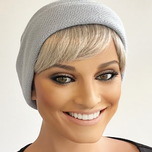 May include: A light gray knit beanie hat. The hat is worn by a person with blonde hair and a black top. The person is smiling, and the image is well-lit.