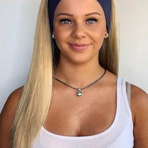 May include: A navy blue headband worn across the forehead. A silver pendant necklace with a round, white pearl-like center. The person is wearing a white tank top and has long blonde hair.