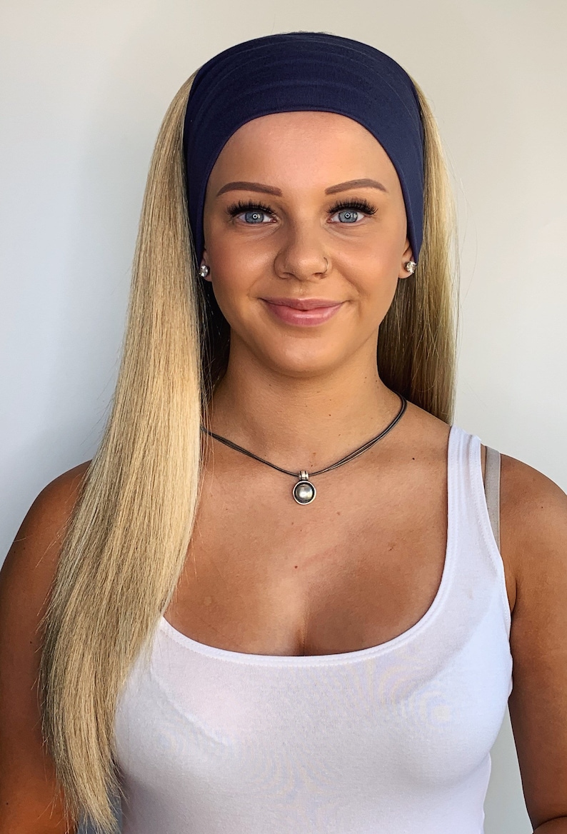 May include: A navy blue headband worn across the forehead. A silver pendant necklace with a round, white pearl-like center. The person is wearing a white tank top and has long blonde hair.