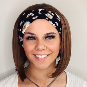 May include: A woman with brown hair wearing a black headband with a white daisy print. She is wearing a white shirt and a silver necklace with a pearl pendant.