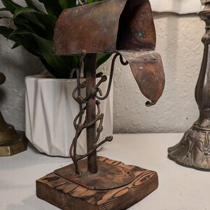 Pode incluir: Uma escultura em miniatura de caixa de correio em metal com um acabamento enferrujado e desgastado. A caixa está montada num poste central adornado com detalhes metálicos retorcidos em forma de videira, sobre uma base quadrada de madeira escura.