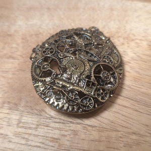 Puede incluir: Broche de estilo steampunk de color bronce. El diseño circular presenta intrincados engranajes, ruedas y elementos mecánicos, creando una estética vintage e industrial. La broche está sobre una superficie de madera.