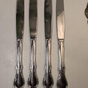 May include: Four silver-toned stainless steel dinner knives arranged vertically. Each knife features a smooth, polished blade and a decorative, ornate handle with embossed scrollwork details at the base and neck of the handle.