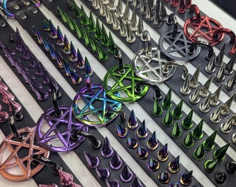 May include: A collection of black leather collars featuring rows of sharp metal spikes and a central pentagram charm. The spikes and charms come in various metallic finishes including iridescent, green, purple, silver, and red, with metal buckles.