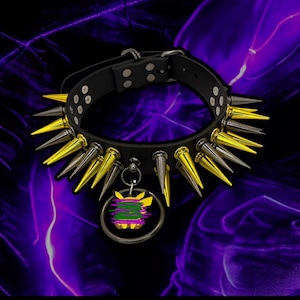 Puede incluir: Un collar de cuero negro con púas doradas y plateadas. El collar tiene un dije adjunto, que presenta un personaje de dibujos animados amarillo con un diseño morado y verde.
