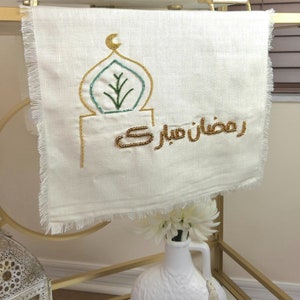 May include: A white linen tea towel with gold and green embroidery. The embroidery depicts a mosque with a crescent moon and a tree. The text "Ramadan Mubarak" is embroidered in gold.