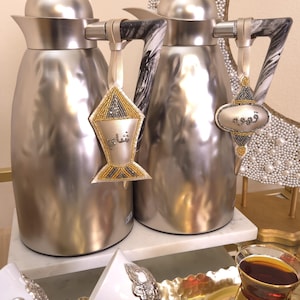 Könnte beinhalten: Zwei silberne Isolierbehälter mit schwarz-weißen Marmorgriffen. Jeder Behälter hat ein dekoratives Etikett mit arabischer Schrift. Ein goldener Tablett enthält ein Glas Tee und zwei verzierte weiße Tassen.