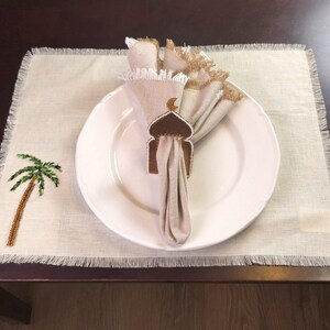 Op de afbeelding: Een wit keramiek bord met een bruine geborduurde servetring in de vorm van een moskee met een halve maan bovenop. De servetring ligt op een beige linnen placemat met een groene geborduurde palmboom.