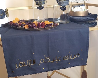 May include: A gold metal serving cart with a black tablecloth featuring the text "مبارك عليكم الشهر" in gold embroidery. The cart is decorated with two clear glass decanters with black labels, a plate of dates and apricots, and a set of small glass cups on a tray.