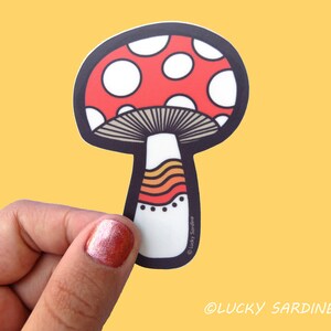 May include: A red and white mushroom sticker with a brown stem and orange and yellow stripes. The sticker has a black outline and the text "&copy; Lucky Sardine" at the bottom.