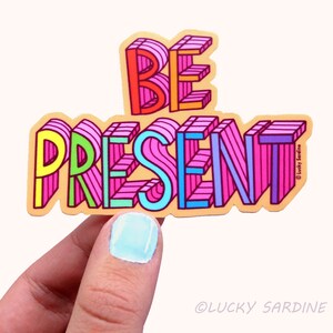 Peut inclure: Un autocollant color&eacute; avec les mots "BE PRESENT" en style 3D. Les lettres sont soulign&eacute;es de noir et remplies de diff&eacute;rentes couleurs, dont le rose, le bleu, le jaune et le vert. L'autocollant est sur un fond brun clair.