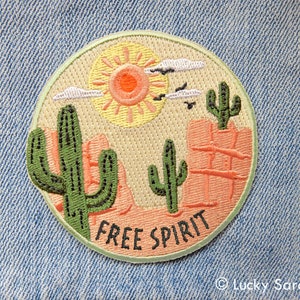 May include: A round embroidered patch with a desert scene. The patch features a yellow sun, two green cacti, and a pink rock formation. The text "FREE SPIRIT" is embroidered in black on the bottom of the patch.
