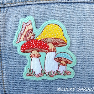May include: Embroidered patch featuring a whimsical design of colorful mushrooms and a butterfly. The patch includes a red mushroom with white spots, a yellow mushroom with white dots, and a blue mushroom. The patch is outlined in a light green color.