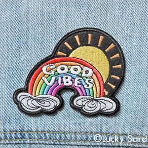 May include: A rainbow with the words "GOOD VIBES" embroidered on it, with a yellow sun behind it. The rainbow is on a denim background.