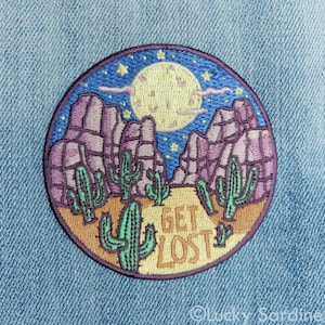 May include: A round embroidered patch with a desert scene. The patch features a full moon, cacti, mountains, and stars. The text "GET LOST" is embroidered in the center of the patch.