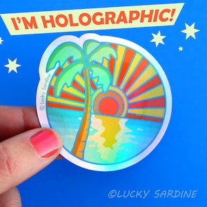 Puede incluir: Una pegatina hologr&aacute;fica con una palmera y una puesta de sol sobre el oc&eacute;ano. La pegatina es redonda y tiene un borde blanco. El texto "I'm Holographic!" est&aacute; escrito encima de la pegatina. El texto "&copy; Lucky Sardine" est&aacute; escrito en la pegatina.