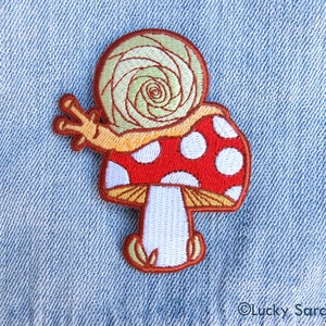 May include: Embroidered iron-on patch featuring a red and white mushroom with a snail on top. The snail is green and yellow and has a spiral shell.