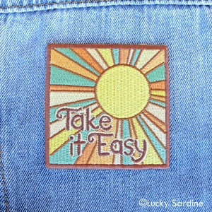 May include: Embroidered iron-on patch with a retro sunburst design in shades of orange, yellow, and green. The text "Take it Easy" is embroidered in brown on the patch.