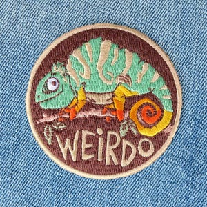 May include: Embroidered patch featuring a green chameleon on a branch, with orange and yellow accents. The patch is circular, with a brown border and the word "WEIRDO" in tan lettering. The patch is set against a blue denim background.
