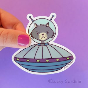 May include: A gray cat wearing a space helmet is piloting a blue and purple flying saucer. The sticker has the text "Lucky Sardine" on the bottom right.