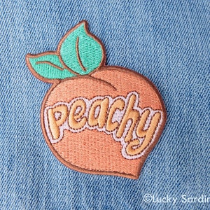 May include: Peach-shaped embroidered patch with the word "peachy" in a pink and brown font on a denim background.