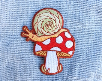 May include: Embroidered iron-on patch featuring a red and white mushroom with a snail on top. The snail is green and yellow and has a spiral shell.