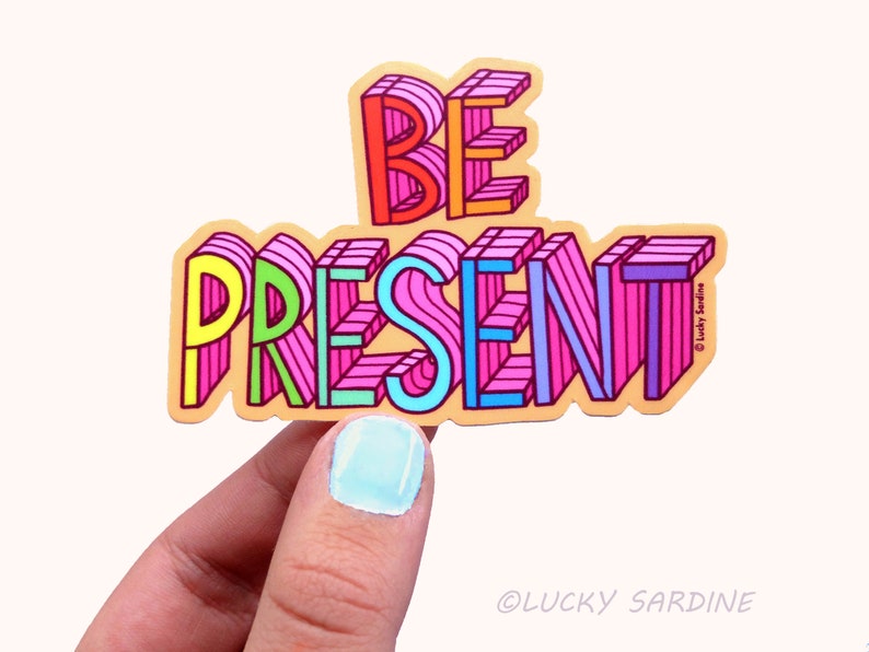 Peut inclure: Un autocollant color&eacute; avec les mots "BE PRESENT" en style 3D. Les lettres sont soulign&eacute;es de noir et remplies de diff&eacute;rentes couleurs, dont le rose, le bleu, le jaune et le vert. L'autocollant est sur un fond brun clair.