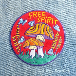 May include: A red, round embroidered patch with the words "Free Spirit" in yellow lettering. The patch features a psychedelic mushroom with orange and yellow stripes, surrounded by green grass and blue butterflies.