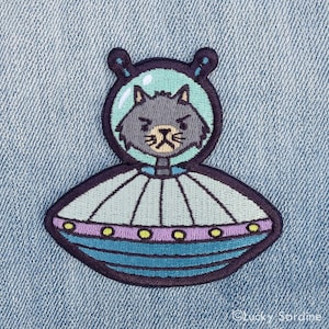 May include: An embroidered iron-on patch of a gray cat in a blue and green spaceship. The cat has a grumpy expression and is looking directly at the viewer.