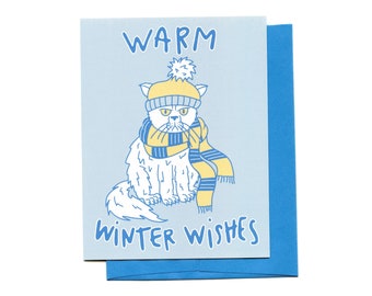 May include: A light blue greeting card with a cartoon cat wearing a yellow hat and scarf. The card reads "WARM WINTER WISHES" in blue lettering. A blue envelope is behind the card. The artist's name, "LUCKY SARDINE", is at the bottom.