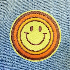 May include: Embroidered patch featuring a retro smiley face design. The patch has a yellow center with brown eyes and a curved smile. The smiley face is surrounded by concentric rings of orange, yellow, and brown. The patch is set against a blue denim background.
