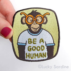 Pu&ograve; includere: Toppa ricamata termoadesiva con una scimmia dei cartoni animati che indossa occhiali rotondi arancioni e una maglietta bianca con il testo "Be a good human".