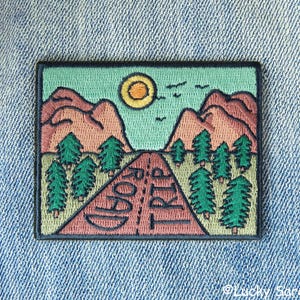 May include: Embroidered patch with a mountain scene, a sun, and the words "Good Trip" on a winding road. The patch is surrounded by a black border.