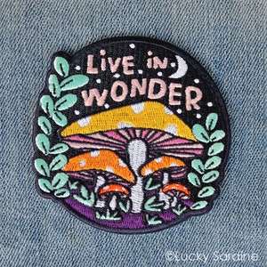 May include: Embroidered patch with a black background, white polka dots, and the text "Live in Wonder". The patch features three colorful mushrooms with white spots and green leaves around the edge.