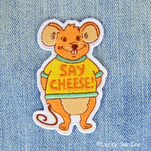 May include: Embroidered patch featuring a cartoon mouse in a yellow shirt with the text "SAY CHEESE!" in orange. The mouse is orange with tan ears and a white outline, set against a blue denim background.
